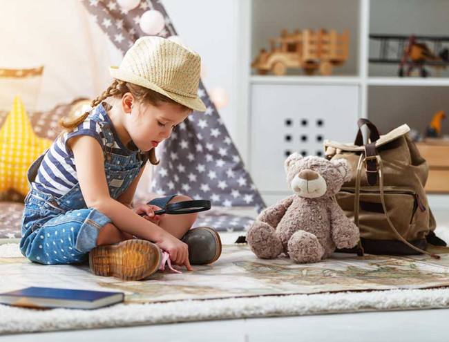 Kleines Mädchen stzt mit Teddybär und Rucksack auf einer Landkarte und sucht mit einer Lupe nach dem Reiseziel.