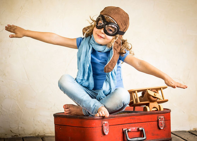Kind mit Fliegerbrille sitzt mit ausgestreckten Armen auf einem Koffer und tut als wäre es ein Flugzeug.